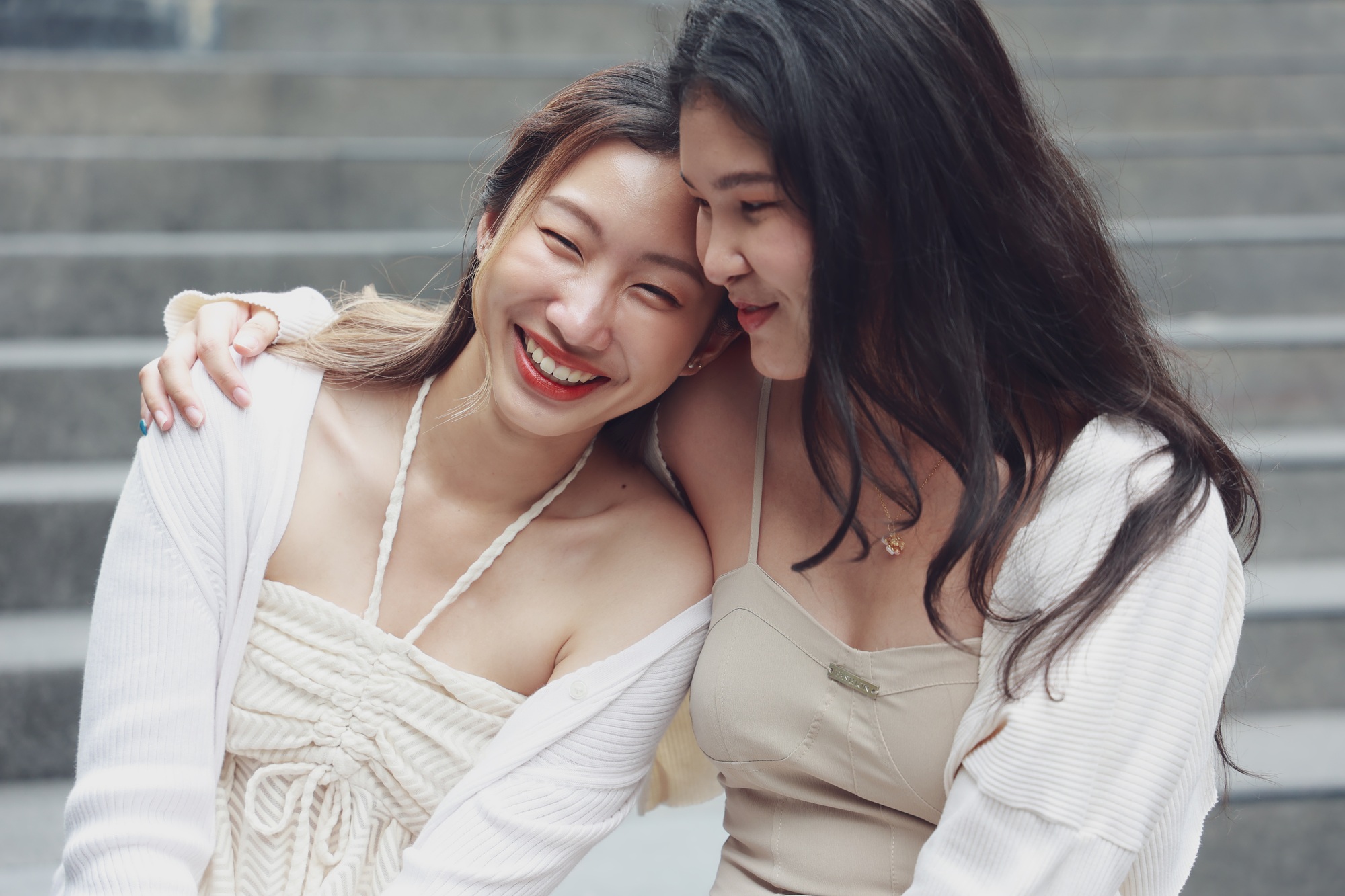 Two joyful women sitting closely and laughing together on steps, showcasing friendship and happiness