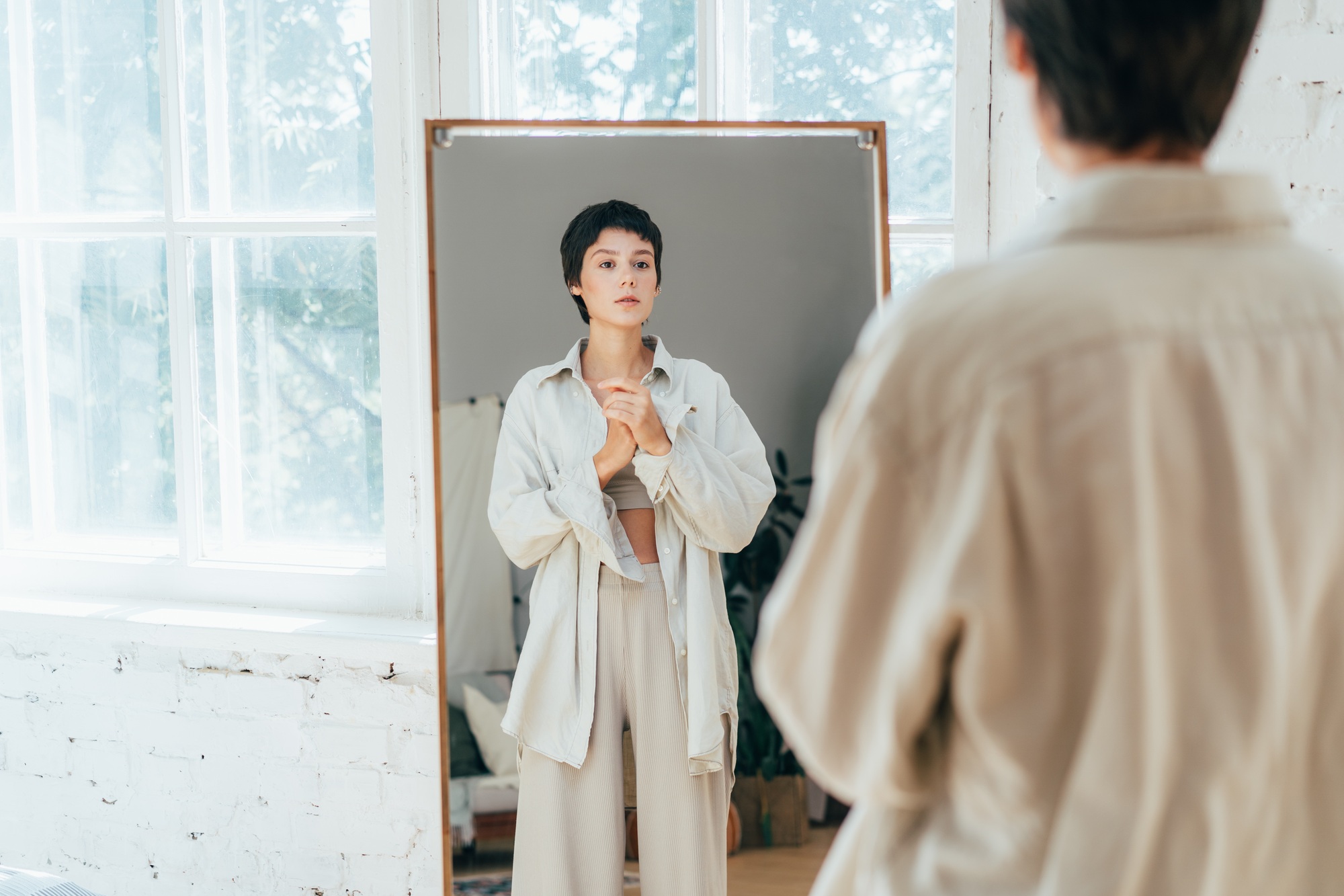 Young modern woman dressed in oversized shirt looking in the mirror at home.