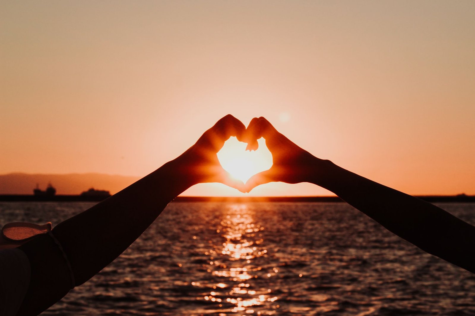 Hands forming a heart shape against an ocean sunset, symbolizing staying open-hearted
